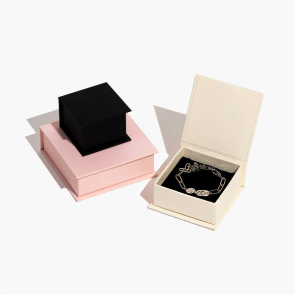 กล่องเครื่องประดับแม่เหล็กแบบกำหนดเองสีดำขนาดเล็กหนึ่งกล่องอยู่ด้านบนของกล่องสีชมพูขนาดใหญ่ และกล่องสีขาวเปิดออกพร้อมสร้อยข้อมือด้านใน