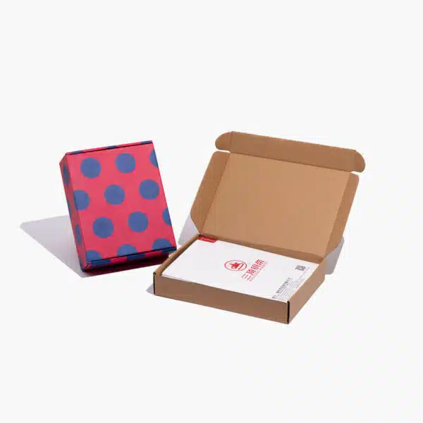 Die rosa Versandschachtel mit blauem Punkt steht neben den individuell gestalteten Kraftpapier-Versandtaschen mit einer Zeitschrift darin
