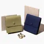 ზეთისხილის მწვანე და ლურჯი მორგებული სამკაულების ჩანთა ღილაკებით დახურული, ცალ-ცალკე დგას ორი კრემისფერი ფიქალის წინააღმდეგ, წინ წყვილი ოქროს საყურეებით.