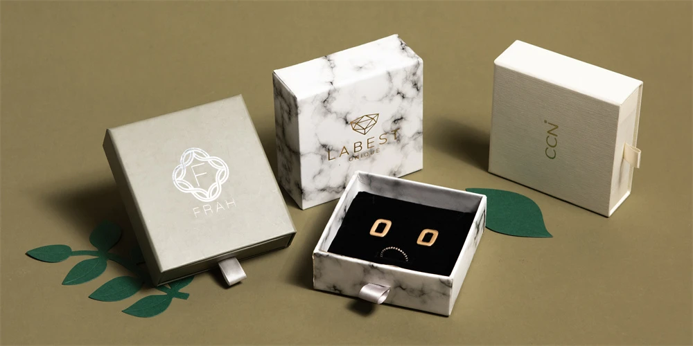 luxury packaging boxes with different brand logos on a green background, one open to show a pair of black earrings inside.