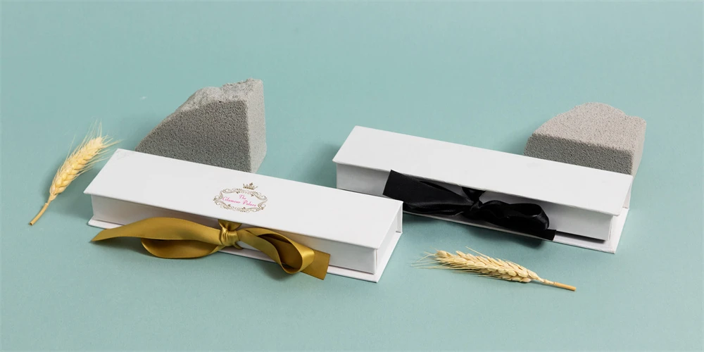 Elegantes cajas de regalo blancas con cintas doradas y negras, acompañadas de tallos de trigo y muestras de piedra, sobre un fondo verde azulado.