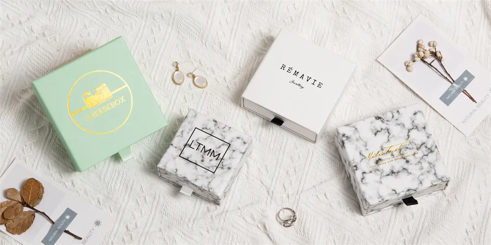 Elegant gift boxes with various brand names are arranged on a textured white surface, accompanied by delicate jewelry and botanical decorations.