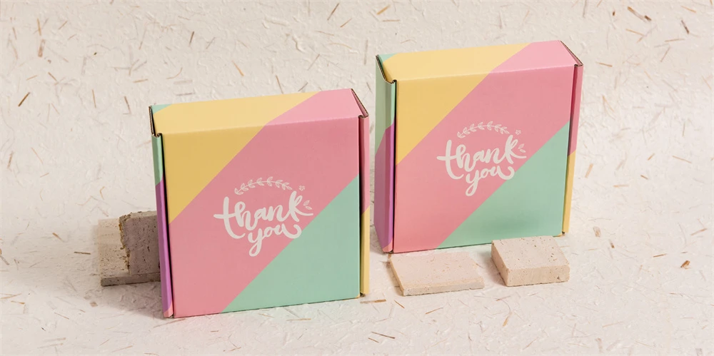Duas caixas de correio "Obrigado" em tons pastéis em tons de rosa, amarelo e verde-azulado, apresentadas em um fundo claro texturizado com amostras de pedra em primeiro plano.