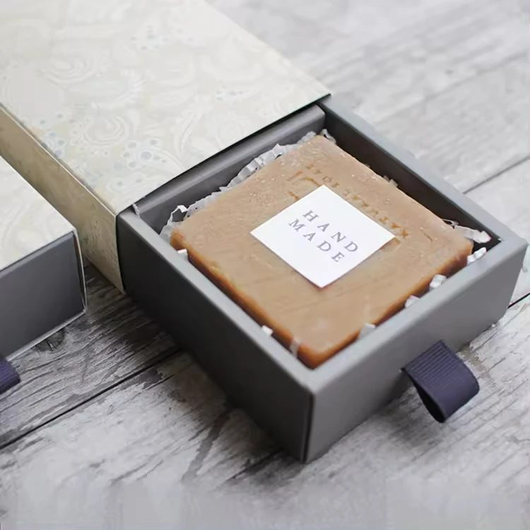 Uma barra de sabonete artesanal com o rótulo "HAND MADE" fica dentro de uma caixa cinza aberta com uma elegante tampa estampada em branco e cinza ao lado, colocada sobre uma superfície de madeira.