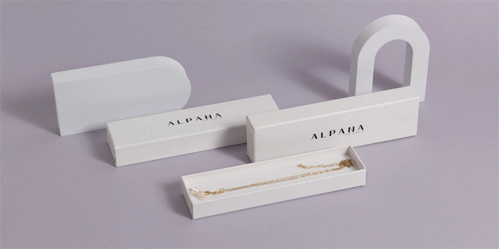 Bele luksuzne embalažne škatle z natisnjenim imenom blagovne znamke, prikazane na vijoličnem ozadju, z eno odprto škatlo, ki razkriva zlato ogrlico.