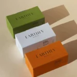 სამი ფერადი ძვირადღირებული საპნის შესაფუთი ყუთი ლამაზად არის დაწყობილი, სადაც წარმოდგენილია თეთრი, მწვანე და ნარინჯისფერი დიზაინი მხიარული ჩრდილებით ღია ფონზე.