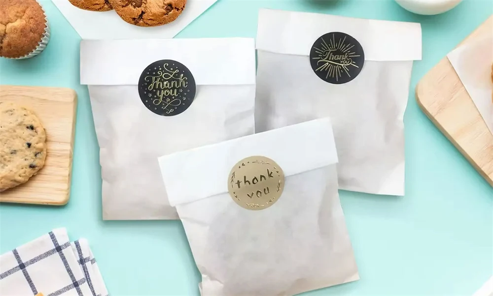 რა არის Glassine Paper-ის ბლოგის ყდა: სამი თეთრი მინის ქაღალდის ჩანთა დალუქული დეკორატიული „გმადლობთ“ სტიკერებით, გარშემორტყმული ფუნთუშებითა და მაფინებით ღია ცისფერ ფონზე.