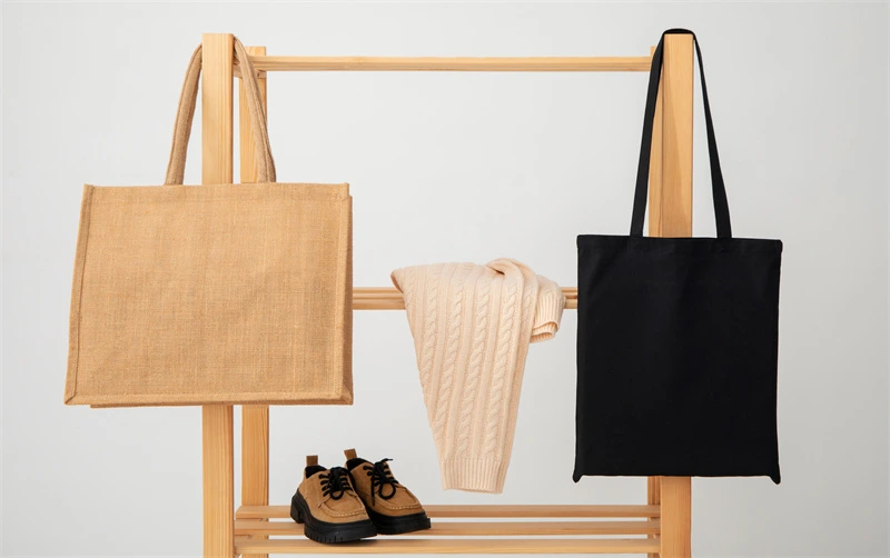 Was ist eine Seitenfaltentasche? Blog-Cover: Zwei Tragetaschen, eine braune und eine schwarze, hängen an einem Holzgestell neben einem beigen Pullover und einem Paar brauner Schuhe.