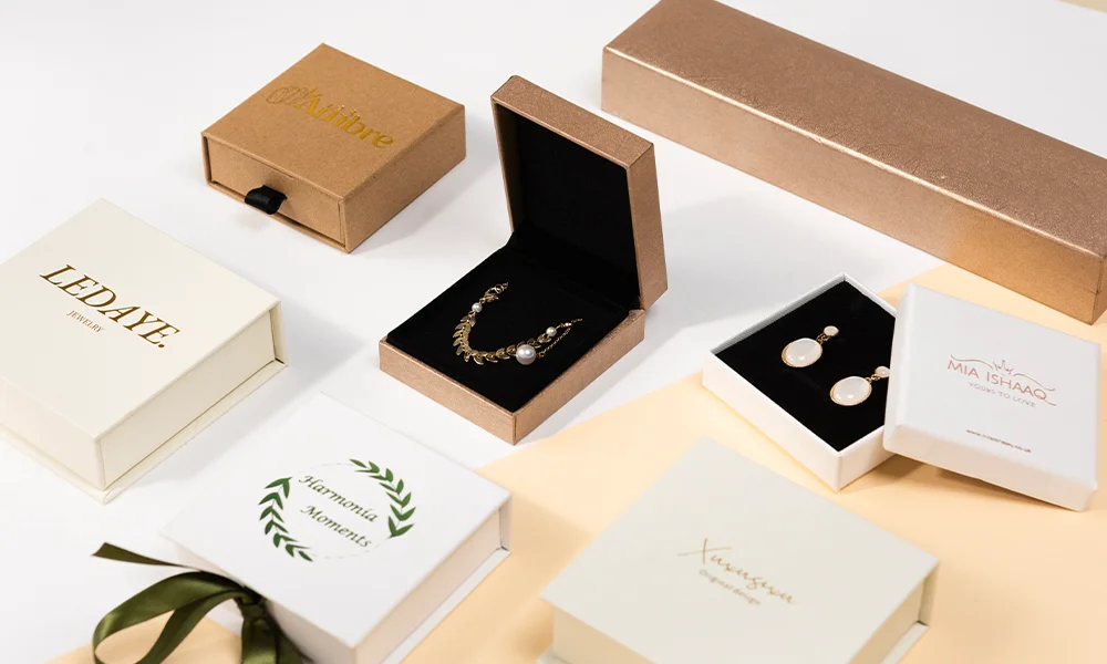 ძვირფასეულობის შეფუთვის იდეების გზამკვლევი საიუველირო ბიზნესის დამწყებთათვის ყდა: მაგიდაზე მიმოფანტულია ყველა სახის საიუველირო ყუთი, ზოგს შიგნით აქვს სამკაულები, ზოგს დახურული.