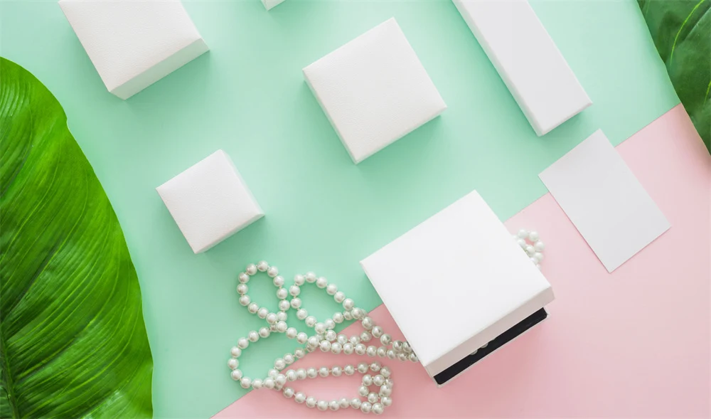 რამდენიმე თეთრი სამკაულის ყუთი მოწესრიგებულად არის მოთავსებული მწვანე და ვარდისფერ ფონზე და მათ გვერდით არის მარგალიტის ყელსაბამი და ფოთლები