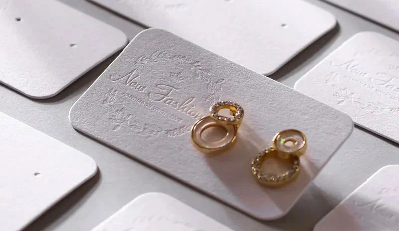 როგორ გააკეთოთ საბაჟო სამკაულები ბარათების ბლოგის გარეკანზე: მორგებული საიუველირო ბარათები გამოსახულია ელეგანტური საყურეებით, რელიეფური "New Fashion" ბრენდით და დეკორატიული ყვავილების დიზაინით.