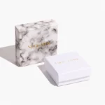 ერთი ფერადი საბაჟო ქაღალდის სამკაულების ყუთი სახურავით, რომელიც დგას თეთრი ყუთის უკან