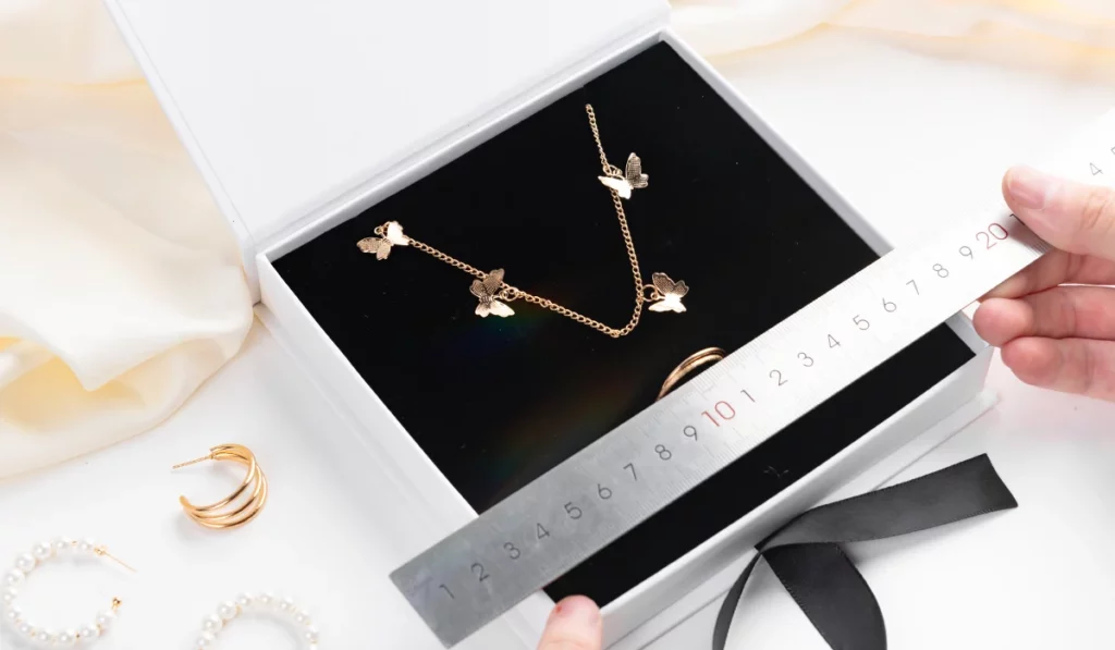 როგორ ზუსტად გავზომოთ სამკაულები, რომ აირჩიოთ სრულყოფილი საიუველირო ყუთის ზომა? ბლოგის ყდა: ოქროს პეპლის თემის ყელსაბამი, რომელიც გამოსახულია შავი ხაზებით სამკაულების ყუთში, რომელსაც სახაზავი ზომავს. ყუთის გარშემო ოქროსა და მარგალიტის საყურეები და ლენტია.