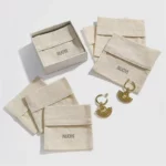 აჩვენეთ მორგებული რბილი ბამბის ტილოს ბრტყელი კონვერტის სამკაულების ჩანთები კრემისფერი ფერით, ასევე არის ყუთი და წყვილი ოქროს საყურეები