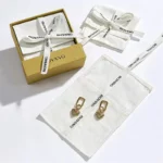 არის სამი თეთრი ბამბის ტილოს კონვერტის სამკაულების ჩანთა სიმებით, ერთი გახსნილია წყვილი ოქროს საყურეებით, ერთი დახურულია, ერთი ყუთშია,