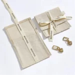 არის ორი ბეჟი მორგებული ბამბის ტილოს კონვერტის სამკაულების ჩანთა სიმებით და წყვილი ოქროს საყურე, ერთი ჩანთა გახსნილია, მეორე დახურულია