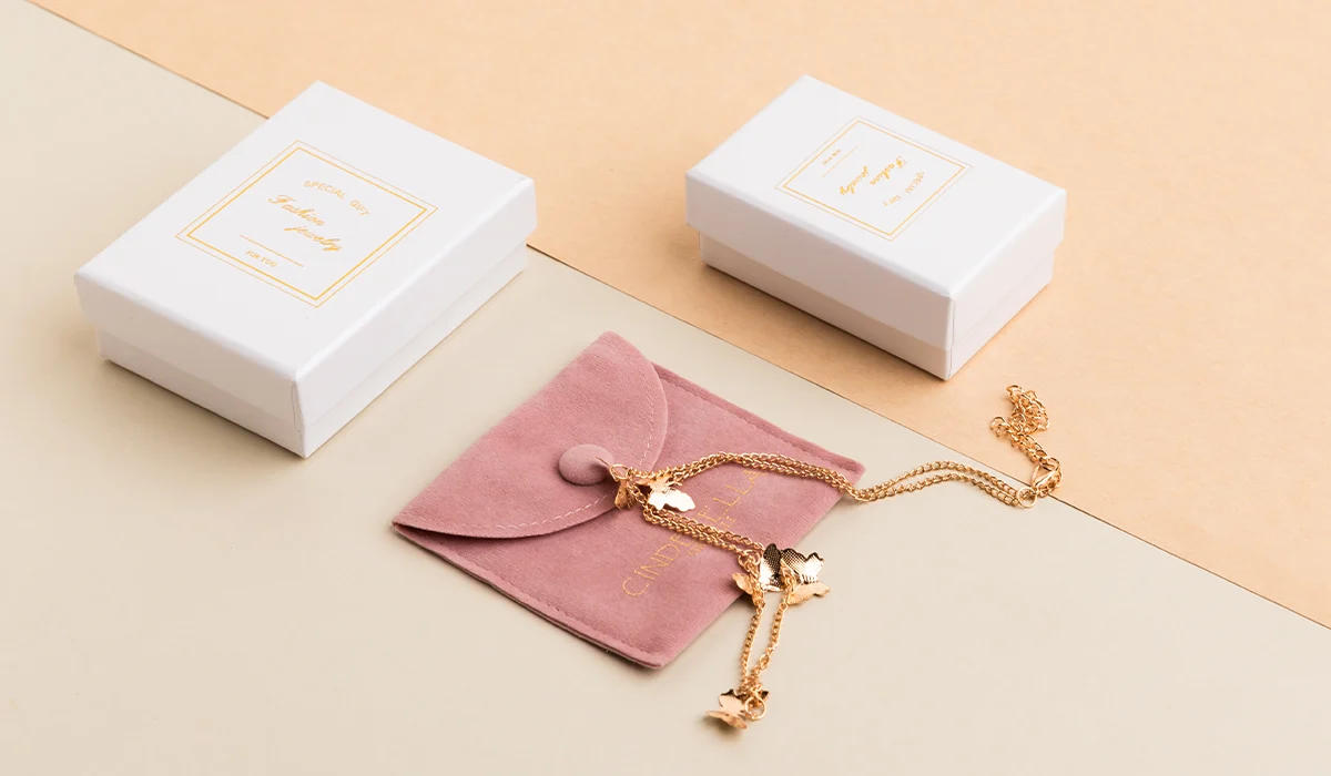 რა არის მორგებული სამკაულების შეფუთვა? ბლოგის ყდა: ელეგანტური თეთრი სამკაულების ყუთები და ვარდისფერი ზამშის ჩანთა, რომელზეც გამოსახულია ოქროს ყელსაბამი პეპლების ხიბლით ნეიტრალურ ფონზე.