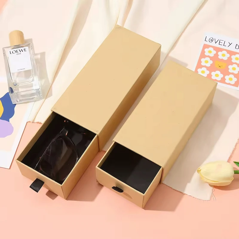 ორი მორგებული კრაფტის ქაღალდის ყუთი გასაშლელი უჯრით, ერთს უჭირავს მზის სათვალე. ფონზე მოჩანს სუნამოს ბოთლი და პატარა ყვავილოვანი ბარათი, ღია ვარდისფერ ზედაპირზე.