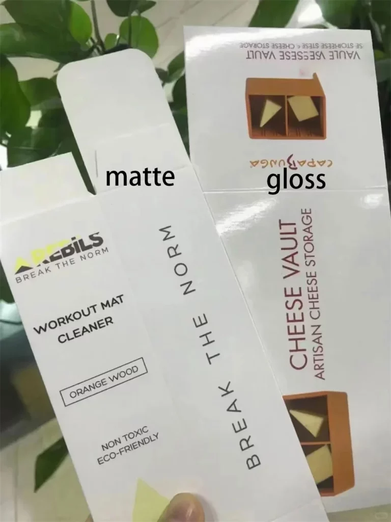 Mattes vs. glänzendes Laminat: Welche Verpackungsoberfläche passt am besten zu Ihrer Marke? 7 Eine Hand hält zwei aufklappbare, individuell gestaltete Markenkartons mit matter und glänzender Laminierung separat.